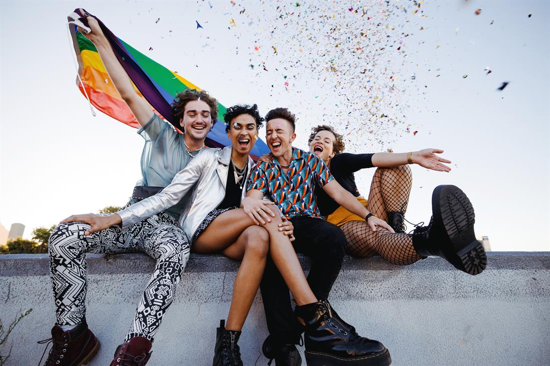Eine Gruppe aus vier Personen unterschiedlichen Geschlechts sitzen auf einer Mauer, im Hintergrund der Himmel. Sie sehen glücklich aus und lachen. Die Person ganz links hält eine Regenbogenflagge in die Luft und die Person ganz rechts wirft Konfetti.