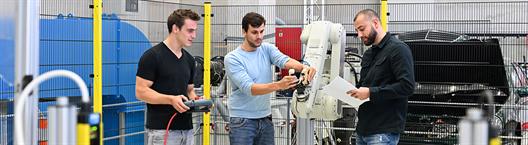 Drei junge Männer in der Maschinenhalle der Hochschule Düsseldorf. Sie untersuchen einen Roboterarm.