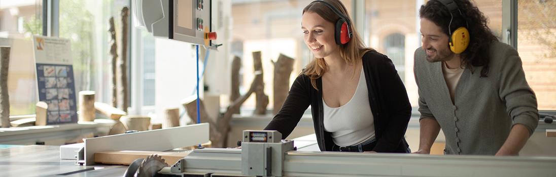 Zwei Studierende bedienen eine elektrische Holzsäge. Sie tragen dabei einen Gehörschutz.