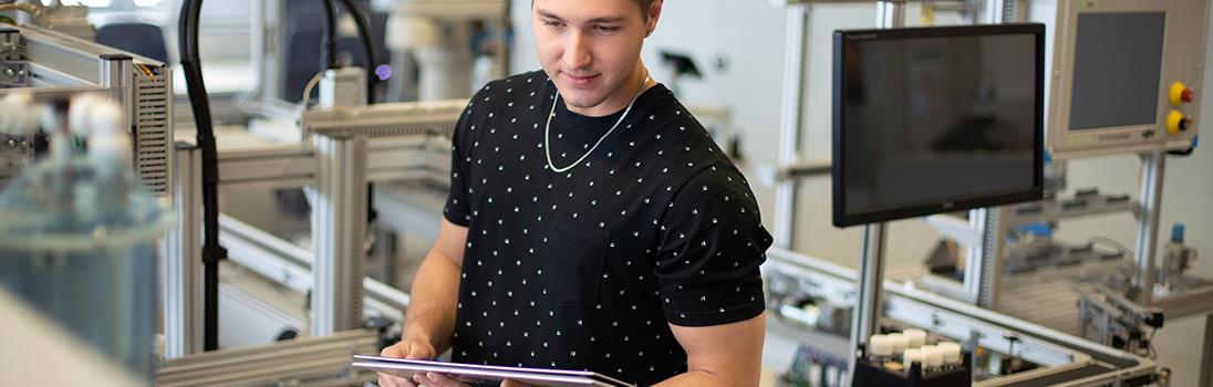 Ein Student steht in einer technischen Anlage und hält ein Tablet in der Hand.