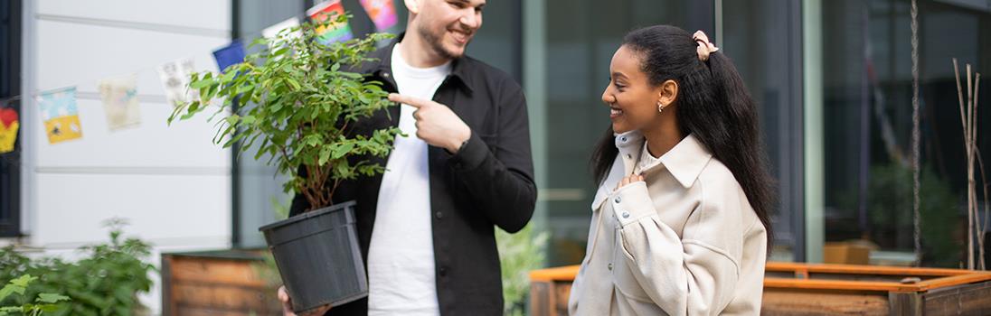 Ein weißer Studierender und eine farbige Studierende stehen zusammen im Urban Garden der Hochschule. Er hält eine Pflanze im Arm und weist mit dem Zeigefinger nach links.