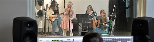 Blick in ein Tonstudio. Hinter der Scheibe sieht man vier junge Frauen, die als Band zusammen musizieren. Im Vordergrund vor der Scheibe sieht man einen Tontechniker.