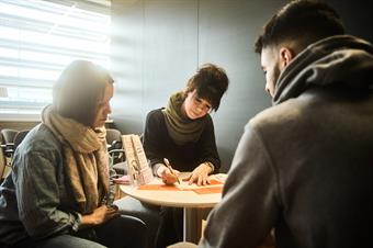 Drei Personen sitzen um einen runden Tisch, auf dem Zettel und Broschüren liegen. Eine Person schreibt etwas auf einen der Zettel.